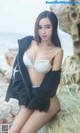 A woman in a white bikini sitting on a rock by the ocean.